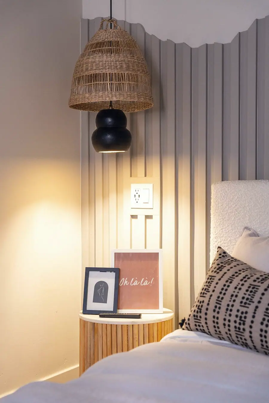 Midway bedside detail with fluted nightstand, woven pendant, and Oh là là art