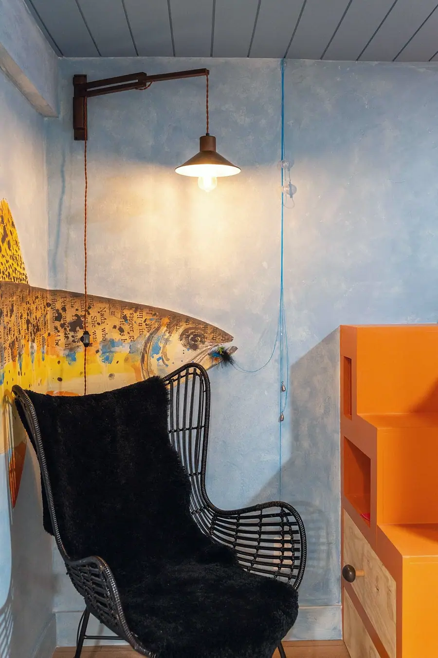 Midway fish mural corner with black rattan chair, swing-arm pendant, and blue plaster wall
